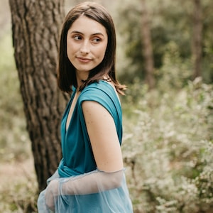 Könnte beinhalten: Eine Frau in einem blaugrünen Kleid mit durchsichtigen blauen Ärmeln steht in einer Waldlandschaft.