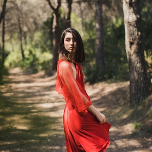 Könnte beinhalten: Eine Frau in einem roten Kleid steht in einer Waldlandschaft. Das Kleid hat einen fließenden Rock und durchsichtige Ärmel. Die Frau schaut in die Kamera.
