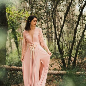 Könnte beinhalten: Eine Frau in einem langen, fließenden rosa Kleid mit einer Spitzenüberlage steht in einem Wald. Das Kleid hat einen tiefen V-Ausschnitt und eine Schärpe, die in der Taille gebunden ist. Die Frau schaut auf den Boden.