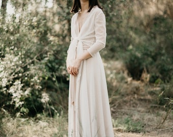 Könnte beinhalten: Eine Frau in einem weißen, gepunkteten Brautkleid mit langem, fließendem Rock und V-Ausschnitt. Sie steht in einer Waldlandschaft mit grünen Bäumen und Blättern.
