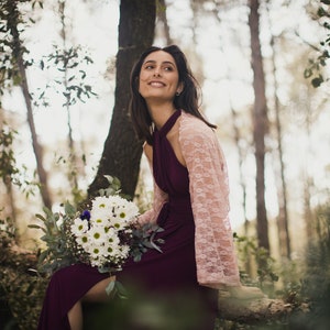 Könnte beinhalten: Eine Frau in einer Waldkulisse trägt ein burgunderfarbenes Maxikleid mit einem rosafarbenen Spitzen-Schal. Sie hält einen Strauß weißer Gänseblümchen und Grünzeug.
