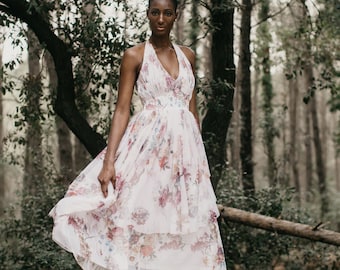 May include: A woman wearing a white floral dress with a halter neckline and a tiered skirt. She is standing in a forest setting.
