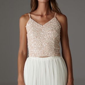 Puede incluir: Una mujer lleva una falda de tul blanca y una blusa de lentejuelas color melocotón. La blusa tiene un escote en V y tirantes finos.