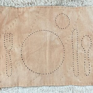 Rectangular cotton placemat for a child. The placemat is dyed with onion skins for a light orange color. The outlines of plate, fork, knife, spoon, and cup are stitched onto the mat in black. The whole mat is quilted with light brown thread.