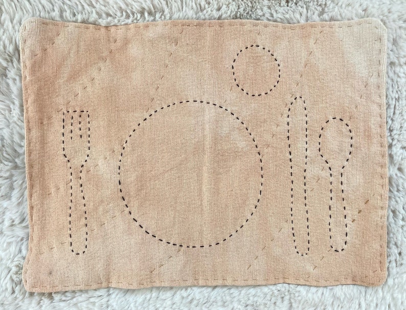 Rectangular cotton placemat for a child. The placemat is dyed with onion skins for a light orange color. The outlines of plate, fork, knife, spoon, and cup are stitched onto the mat in black. The whole mat is quilted with light brown thread.