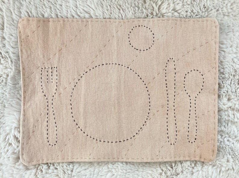 Rectangular cotton placemat for a child. The placemat is naturally dyed with avocado for a light mauve color. The outlines of plate, fork, knife, spoon, and cup are stitched onto the mat in black. The whole mat is quilted with light brown thread.