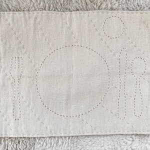 Rectangular cotton placemat for a child. The placemat is an undyed white color. The outlines of plate, fork, knife, spoon, and cup are stitched onto the mat in light brown. The whole mat is quilted with gray thread.