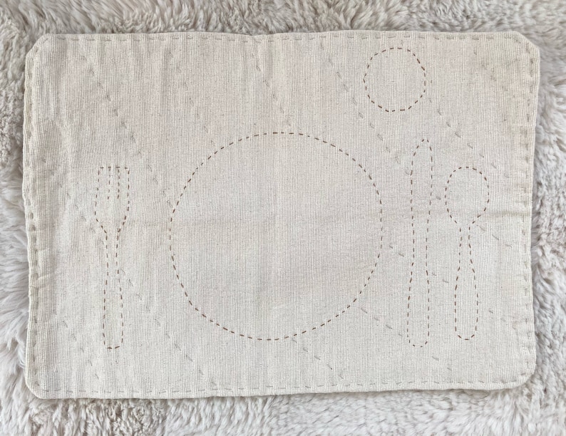 Rectangular cotton placemat for a child. The placemat is an undyed white color. The outlines of plate, fork, knife, spoon, and cup are stitched onto the mat in light brown. The whole mat is quilted with gray thread.