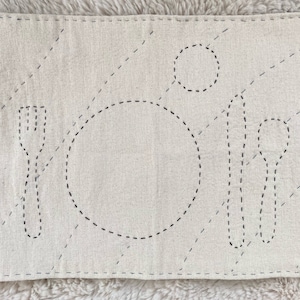 Rectangular cotton placemat for a child. The placemat is undyed natural white. The outlines of plate, fork, knife, spoon, and cup are stitched onto the mat in black. The whole mat is quilted with blue thread.