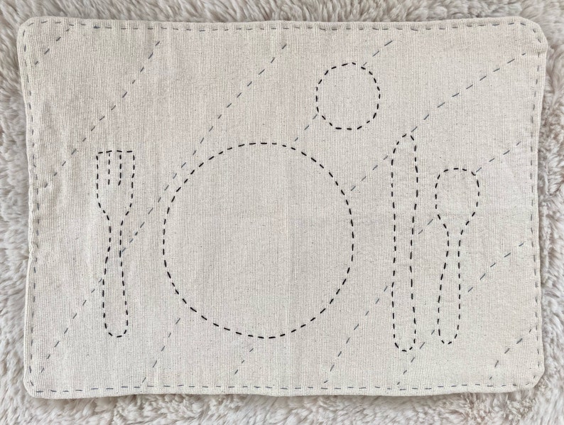 Rectangular cotton placemat for a child. The placemat is undyed natural white. The outlines of plate, fork, knife, spoon, and cup are stitched onto the mat in black. The whole mat is quilted with blue thread.