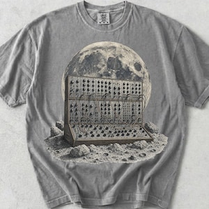 Puede incluir: Una camiseta gris de manga corta con un estampado gráfico de un sintetizador modular sobre una superficie lunar con cráteres frente a una luna grande y detallada. La camiseta tiene cuello redondo y una etiqueta de marca visible en el cuello.