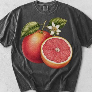 Puede incluir: Una camiseta gris carb&oacute;n de manga corta con una ilustraci&oacute;n bot&aacute;nica de un pomelo entero, un pomelo cortado por la mitad con interior rosa vibrante y una peque&ntilde;a flor de c&iacute;trico blanca con hojas verdes centrada en la parte delantera.