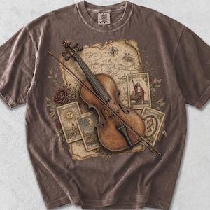 May include: A brown short-sleeve t-shirt featuring a graphic print of a violin with a bow, layered over a vintage-style map and three tarot cards. The design includes floral accents and the word ESPRESSO printed in black below the shirt.