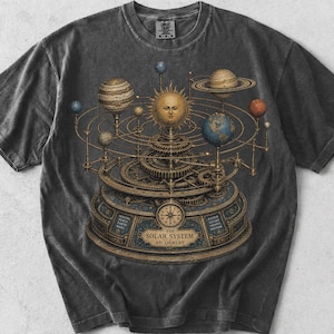 May include: A charcoal gray short-sleeve t-shirt featuring a detailed graphic of a mechanical solar system orrery. The design includes a central sun, orbiting planets, and intricate gears, with the text THE SOLAR SYSTEM AN ORRERY printed below.