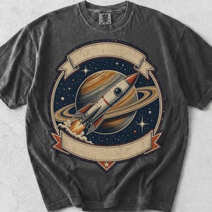 May include: A charcoal gray short-sleeve t-shirt featuring a circular graphic of a rocket ship launching in front of a ringed planet. The design includes a starry space background and two blank beige banners at the top and bottom of the circle.