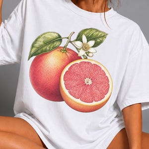 Puede incluir: Una camiseta blanca de manga corta con un gran estampado gr&aacute;fico detallado de un pomelo entero y un pomelo cortado por la mitad con hojas verdes y una peque&ntilde;a flor blanca centrado en la parte delantera de la prenda.