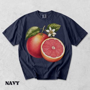 Puede incluir: Una camiseta azul marino de manga corta con una ilustraci&oacute;n bot&aacute;nica de un pomelo entero, un pomelo cortado por la mitad con segmentos rosados visibles, hojas verdes y una flor blanca centrada en el pecho. Cuello redondo.
