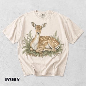 May include: An ivory short-sleeve t-shirt featuring a detailed illustration of a spotted deer resting in a patch of green ferns. The deer is light brown with white spots, and the graphic is centered on the chest of the garment.