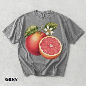 Puede incluir: Una camiseta gris de manga corta con un estampado gr&aacute;fico de un pomelo entero, un pomelo cortado por la mitad con interior rosa y una flor de c&iacute;trico blanca con hojas verdes. La camiseta tiene cuello redondo y una etiqueta de marca visible.