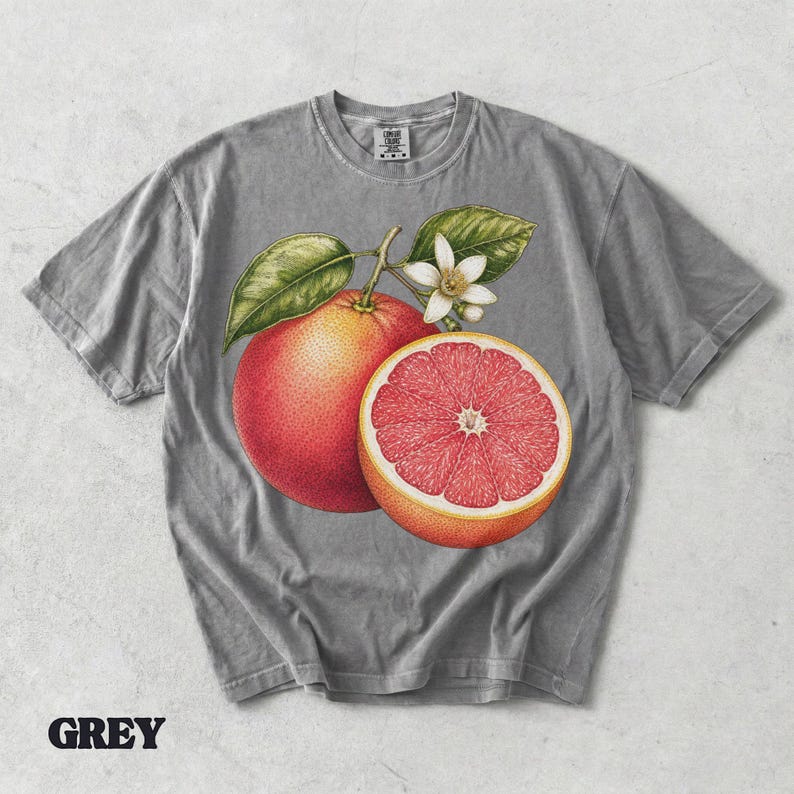 Puede incluir: Una camiseta gris de manga corta con un estampado gr&aacute;fico de un pomelo entero, un pomelo cortado por la mitad con interior rosa y una flor de c&iacute;trico blanca con hojas verdes. La camiseta tiene cuello redondo y una etiqueta de marca visible.