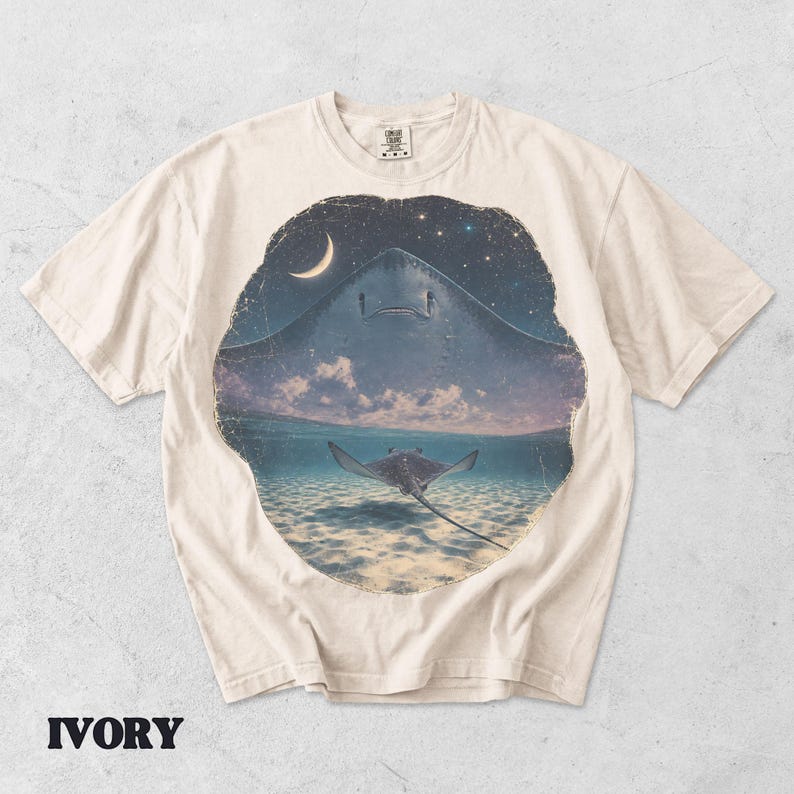 Pode incluir: Uma t-shirt cor de marfim com uma estampa gr&aacute;fica circular de uma raia a nadar em &aacute;guas rasas sob um c&eacute;u noturno com uma lua crescente e estrelas. O design mostra uma grande silhueta de raia no c&eacute;u acima de uma raia menor no oceano.