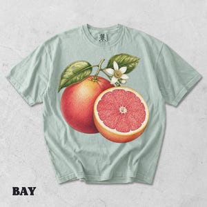 Puede incluir: Una camiseta de color verde salvia claro con una ilustraci&oacute;n bot&aacute;nica detallada de un pomelo entero y un pomelo cortado por la mitad con una flor blanca y hojas verdes. La camiseta tiene cuello redondo y mangas cortas con una etiqueta.