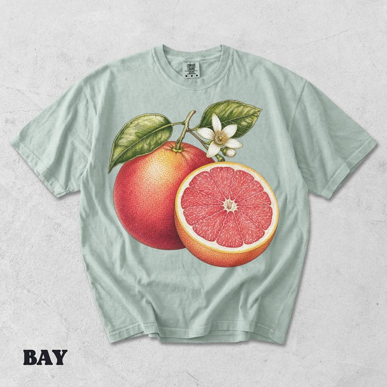 Puede incluir: Una camiseta de color verde salvia claro con una ilustraci&oacute;n bot&aacute;nica detallada de un pomelo entero y un pomelo cortado por la mitad con una flor blanca y hojas verdes. La camiseta tiene cuello redondo y mangas cortas con una etiqueta.