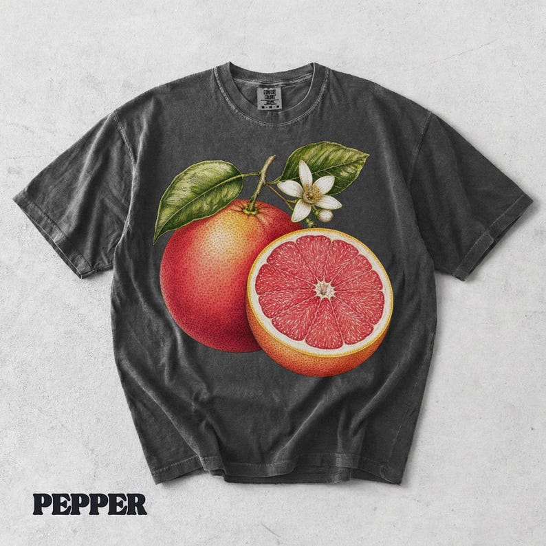 Puede incluir: Una camiseta gris carb&oacute;n de manga corta con una ilustraci&oacute;n bot&aacute;nica de un pomelo entero, un pomelo cortado por la mitad con interior rosa vibrante y una peque&ntilde;a flor de c&iacute;trico blanca con hojas verdes centrada en la parte delantera.