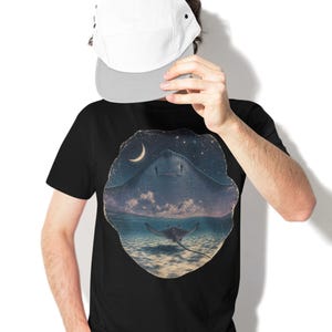 Pode incluir: Uma t-shirt preta com uma estampa gr&aacute;fica circular de uma raia a nadar num oceano escuro sob um c&eacute;u noturno estrelado com uma lua crescente. A t-shirt &eacute; combinada com um bon&eacute; branco com aba cinzenta clara e cal&ccedil;as de cor c&aacute;qui.