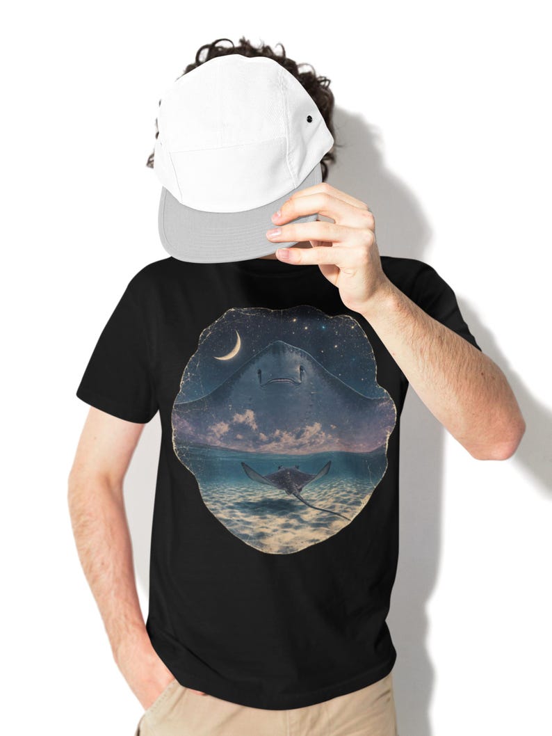 Pode incluir: Uma t-shirt preta com uma estampa gr&aacute;fica circular de uma raia a nadar num oceano escuro sob um c&eacute;u noturno estrelado com uma lua crescente. A t-shirt &eacute; combinada com um bon&eacute; branco com aba cinzenta clara e cal&ccedil;as de cor c&aacute;qui.