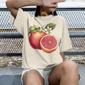 Puede incluir: Una camiseta de manga corta de color crema con un estampado gr&aacute;fico detallado de un pomelo entero y un pomelo cortado por la mitad con hojas verdes y una flor blanca centrado en la parte delantera de la prenda.
