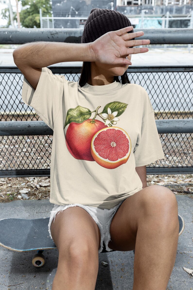 Puede incluir: Una camiseta de manga corta de color crema con un estampado gr&aacute;fico detallado de un pomelo entero y un pomelo cortado por la mitad con hojas verdes y una flor blanca centrado en la parte delantera de la prenda.