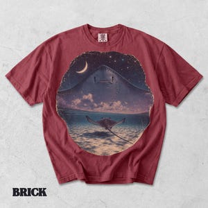 Pode incluir: Uma t-shirt de manga curta cor de tijolo com uma impress&atilde;o gr&aacute;fica circular. O design mostra a silhueta de uma raia grande contra um c&eacute;u noturno estrelado com uma lua crescente, e uma raia menor a nadar em &aacute;gua l&iacute;mpida do oceano.
