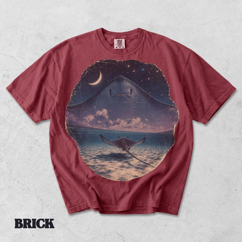 Pode incluir: Uma t-shirt de manga curta cor de tijolo com uma impress&atilde;o gr&aacute;fica circular. O design mostra a silhueta de uma raia grande contra um c&eacute;u noturno estrelado com uma lua crescente, e uma raia menor a nadar em &aacute;gua l&iacute;mpida do oceano.