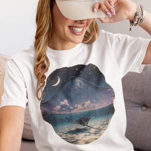 Pode incluir: Uma t-shirt branca de manga curta com uma estampa gr&aacute;fica circular de uma raia a nadar num oceano pouco profundo sob um c&eacute;u noturno com uma lua crescente e estrelas. O design mostra a silhueta de uma raia grande sobre a &aacute;gua.