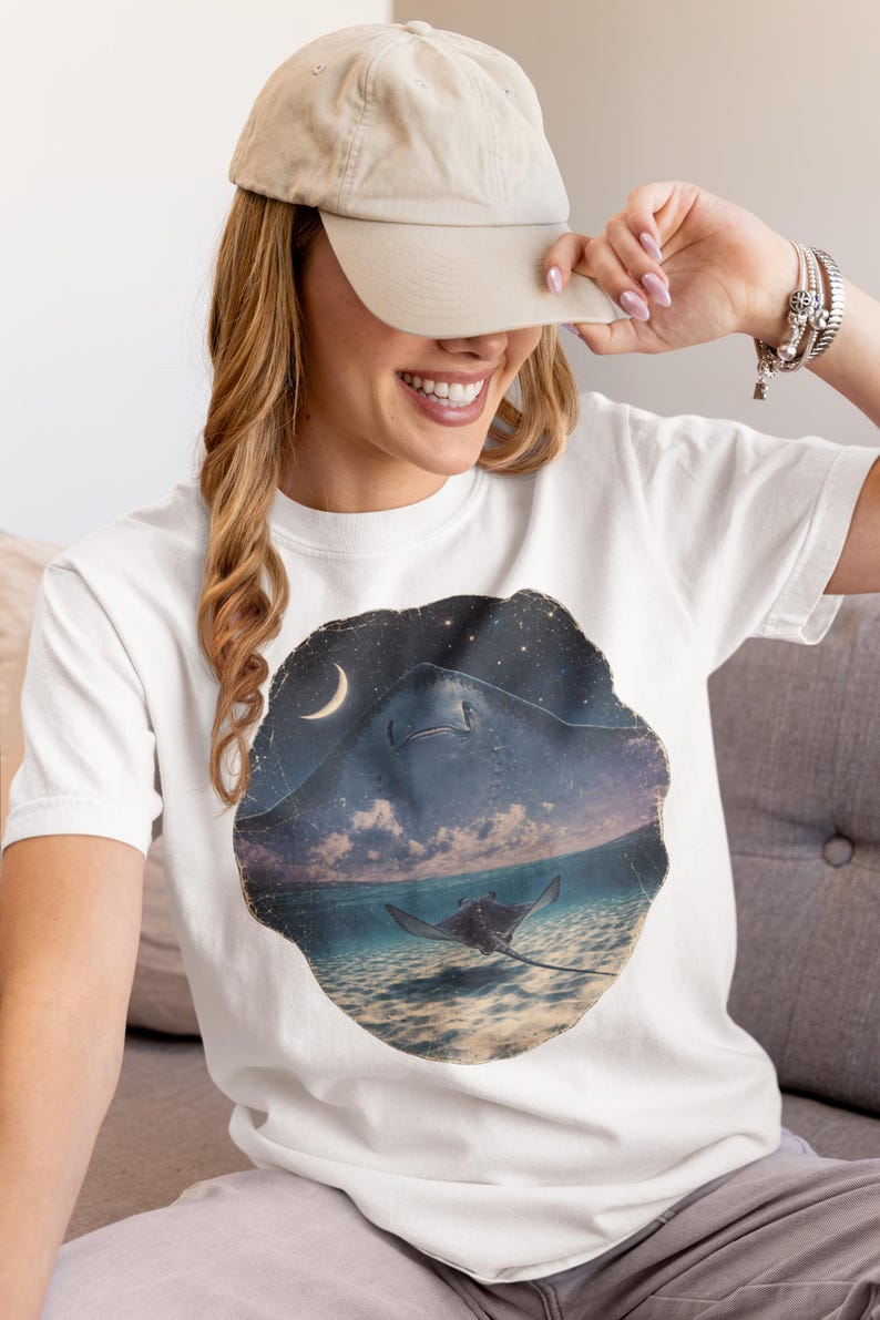 Pode incluir: Uma t-shirt branca de manga curta com uma estampa gr&aacute;fica circular de uma raia a nadar num oceano pouco profundo sob um c&eacute;u noturno com uma lua crescente e estrelas. O design mostra a silhueta de uma raia grande sobre a &aacute;gua.