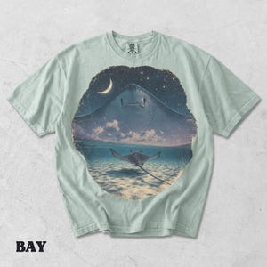 Pode incluir: Uma t-shirt verde s&aacute;lvia claro com uma estampa gr&aacute;fica circular. O design mostra a silhueta de uma grande raia contra um c&eacute;u noturno estrelado com uma lua crescente, que transita para uma raia a nadar em &aacute;guas marinhas claras e rasas.