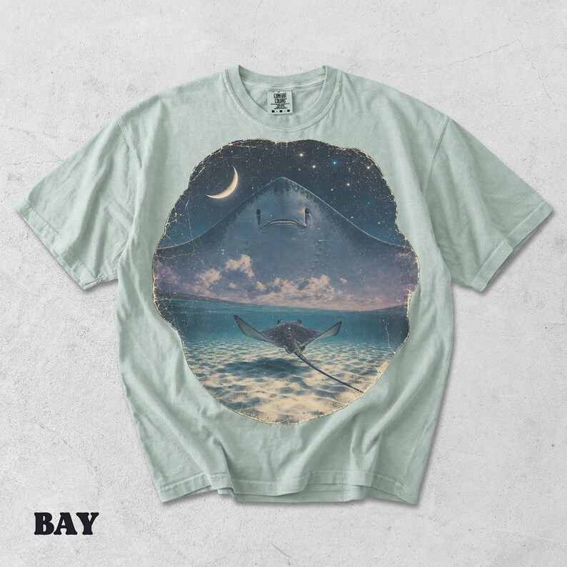 Pode incluir: Uma t-shirt verde s&aacute;lvia claro com uma estampa gr&aacute;fica circular. O design mostra a silhueta de uma grande raia contra um c&eacute;u noturno estrelado com uma lua crescente, que transita para uma raia a nadar em &aacute;guas marinhas claras e rasas.
