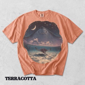 Pode incluir: Uma t-shirt de cor terracota com uma impress&atilde;o gr&aacute;fica circular de uma raia a nadar em &aacute;guas pouco profundas sob um c&eacute;u noturno com uma lua crescente e estrelas. O design mostra a silhueta de uma raia grande sobre uma mais pequena na &aacute;gua.