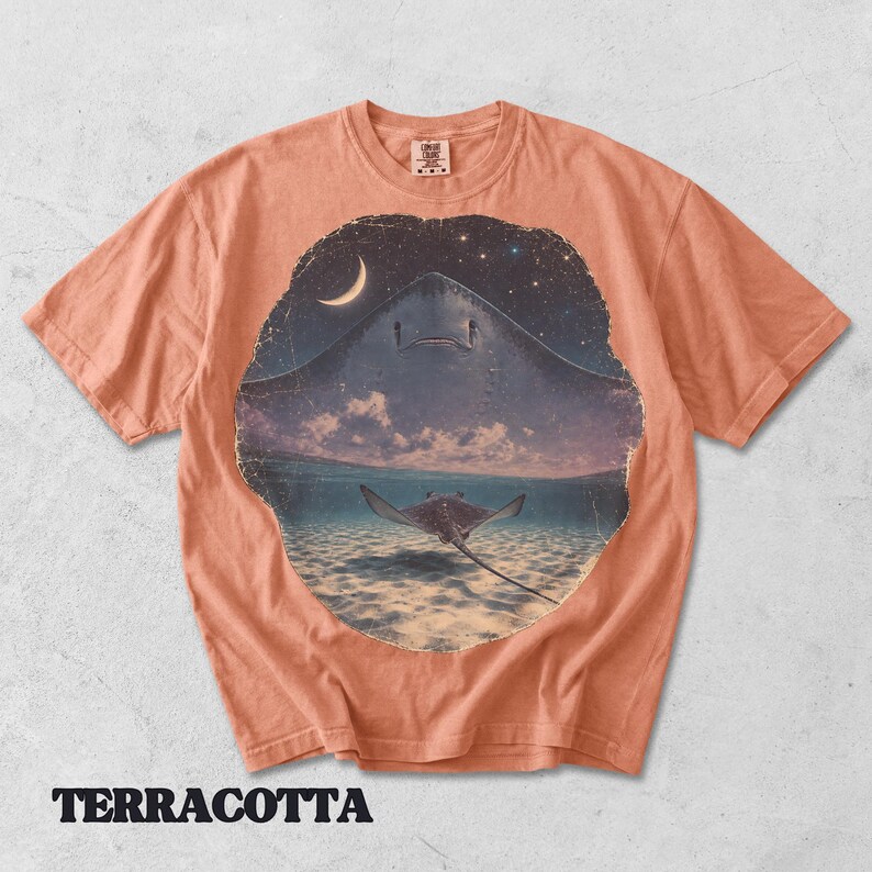 Pode incluir: Uma t-shirt de cor terracota com uma impress&atilde;o gr&aacute;fica circular de uma raia a nadar em &aacute;guas pouco profundas sob um c&eacute;u noturno com uma lua crescente e estrelas. O design mostra a silhueta de uma raia grande sobre uma mais pequena na &aacute;gua.