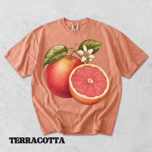 Puede incluir: Una camiseta de color terracota con una ilustraci&oacute;n bot&aacute;nica de un pomelo entero, un pomelo cortado por la mitad con segmentos rosados, hojas verdes y una flor blanca. La camiseta tiene cuello redondo y una etiqueta en el interior del cuello.