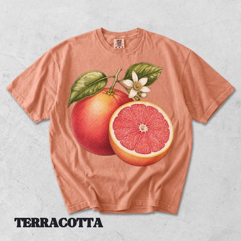 Puede incluir: Una camiseta de color terracota con una ilustraci&oacute;n bot&aacute;nica de un pomelo entero, un pomelo cortado por la mitad con segmentos rosados, hojas verdes y una flor blanca. La camiseta tiene cuello redondo y una etiqueta en el interior del cuello.