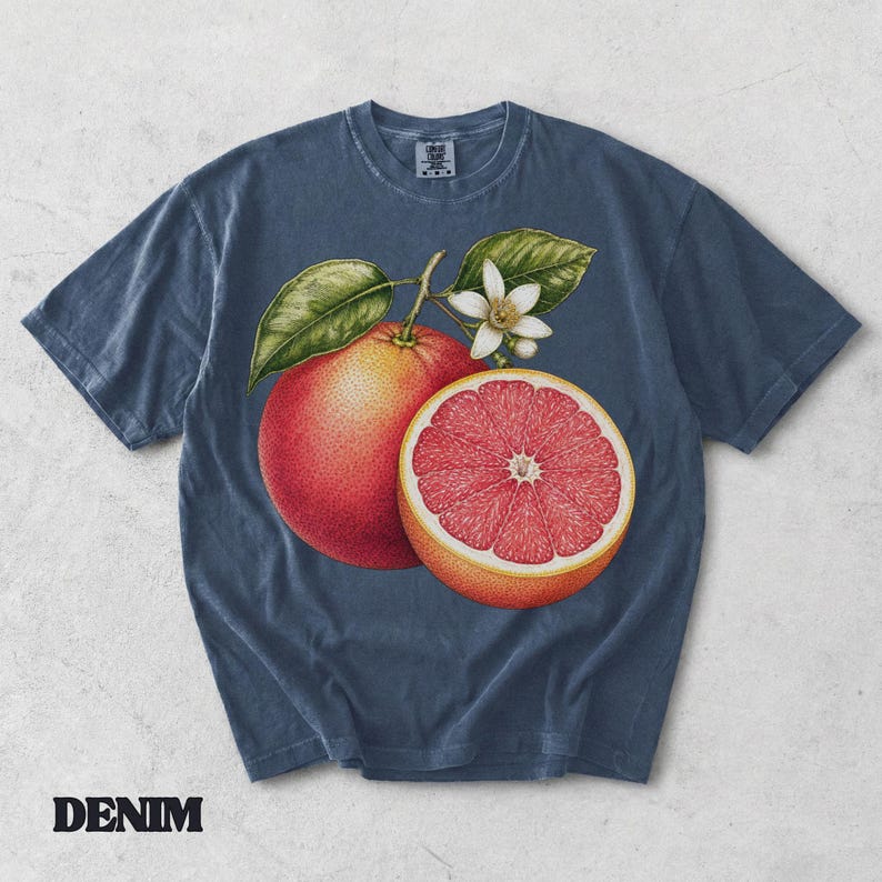 Puede incluir: Una camiseta de manga corta color azul pizarra con una ilustraci&oacute;n gr&aacute;fica de un pomelo entero, un pomelo cortado por la mitad con interior rosa y una flor de c&iacute;trico blanca con hojas verdes en el centro. La camiseta tiene cuello redondo.