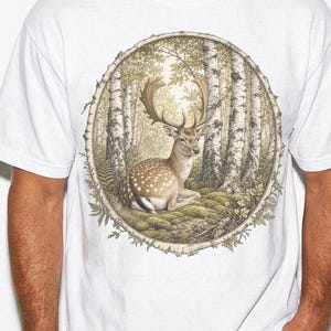 May include: A white short-sleeved t-shirt featuring a circular graphic print of a fallow deer with large antlers resting on a mossy forest floor. The scene is framed by a textured tree bark border and surrounded by tall birch trees in the background.