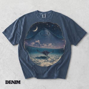 Pode incluir: Uma t-shirt de manga curta em azul ganga com uma grande impress&atilde;o gr&aacute;fica circular. O design mostra uma raia a nadar em &aacute;guas pouco profundas sob um c&eacute;u noturno estrelado com uma lua crescente. A t-shirt tem um decote redondo.