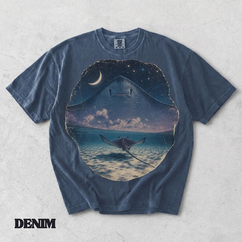 Pode incluir: Uma t-shirt de manga curta em azul ganga com uma grande impress&atilde;o gr&aacute;fica circular. O design mostra uma raia a nadar em &aacute;guas pouco profundas sob um c&eacute;u noturno estrelado com uma lua crescente. A t-shirt tem um decote redondo.