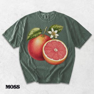 Puede incluir: Una camiseta de manga corta color verde musgo con una ilustraci&oacute;n bot&aacute;nica de un pomelo entero y otro cortado por la mitad, junto a una flor blanca y hojas verdes en el centro. La prenda cuenta con un cuello redondo y acabado vintage.