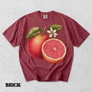 Puede incluir: Una camiseta de manga corta color rojo ladrillo con un estampado gr&aacute;fico de un pomelo entero, un pomelo cortado por la mitad, hojas verdes y una flor blanca. La etiqueta interior del cuello muestra el texto Comfort Colors.