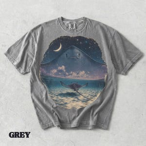 Pode incluir: Uma t-shirt cinzenta de manga curta com uma impress&atilde;o gr&aacute;fica circular de uma raia grande a nadar num oceano l&iacute;mpido sob um c&eacute;u noturno com uma lua crescente e estrelas. O design mostra uma segunda raia mais pequena debaixo de &aacute;gua.