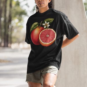 Puede incluir: Una camiseta negra de manga corta con un estampado gr&aacute;fico detallado de un pomelo entero y un pomelo cortado por la mitad con hojas verdes y una peque&ntilde;a flor blanca centrado en la zona del pecho de la prenda.
